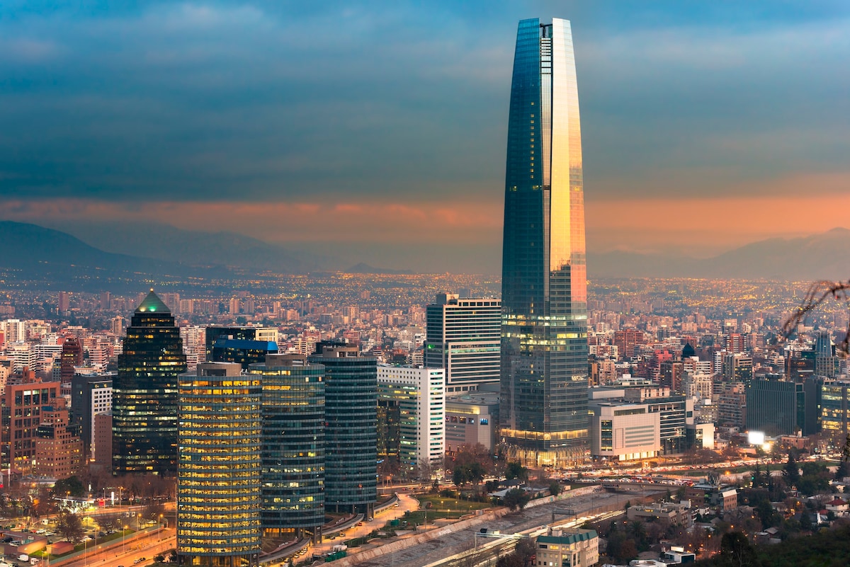 A panoramic view of Santiago's skyline captures tall glass buildings reflecting the evening light. The prominent Costanera Center skyscraper stands out, surrounded by a cityscape that features a mix of modern architecture and distant mountains under a dramatic sky.