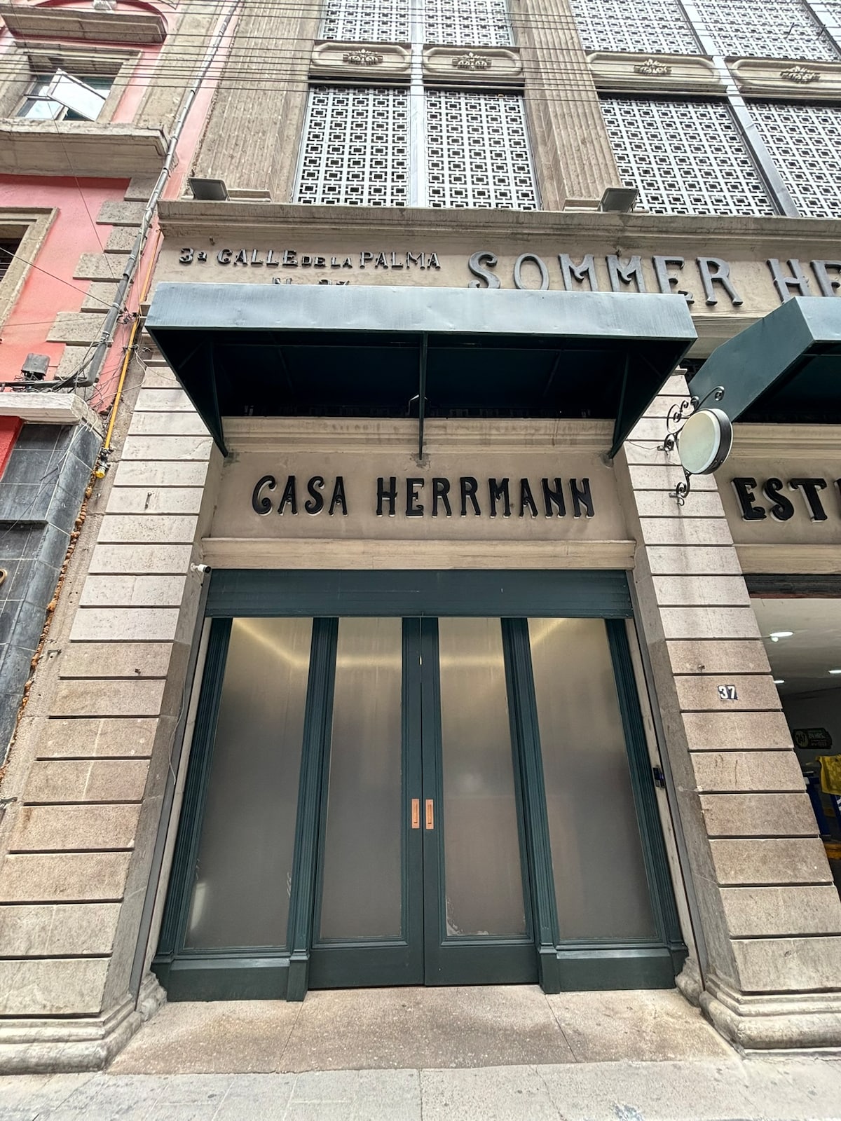 The entrance of Casa Herrmann features a large glass door framed by a dark green overhang. The building's facade displays textured stone walls with decorative details, showcasing an elegant and historic architectural style. Light filters through the glass, highlighting the welcoming entry.