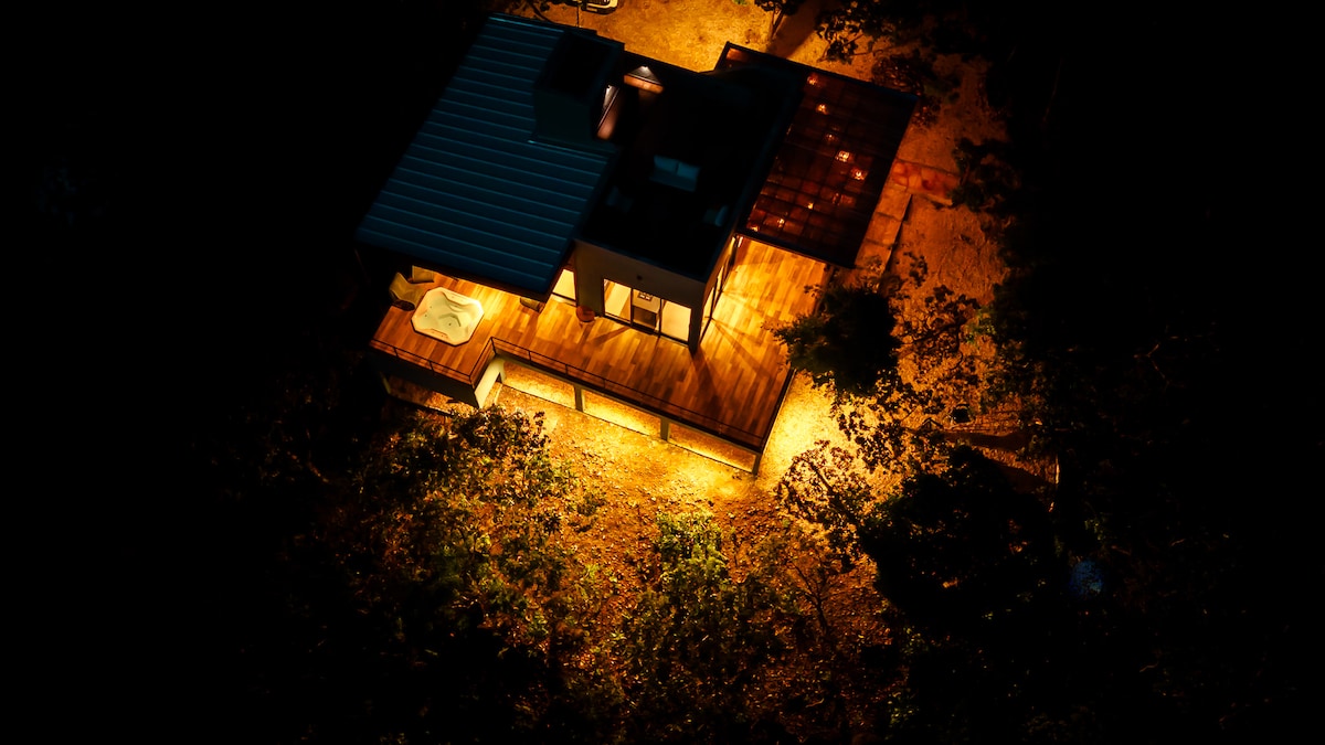 Aerial view of a modern house illuminated at night, surrounded by trees. The house features a spacious wooden deck and a distinctive roof, with warm light highlighting its structure and surrounding pathways. The natural setting offers a serene and secluded atmosphere.