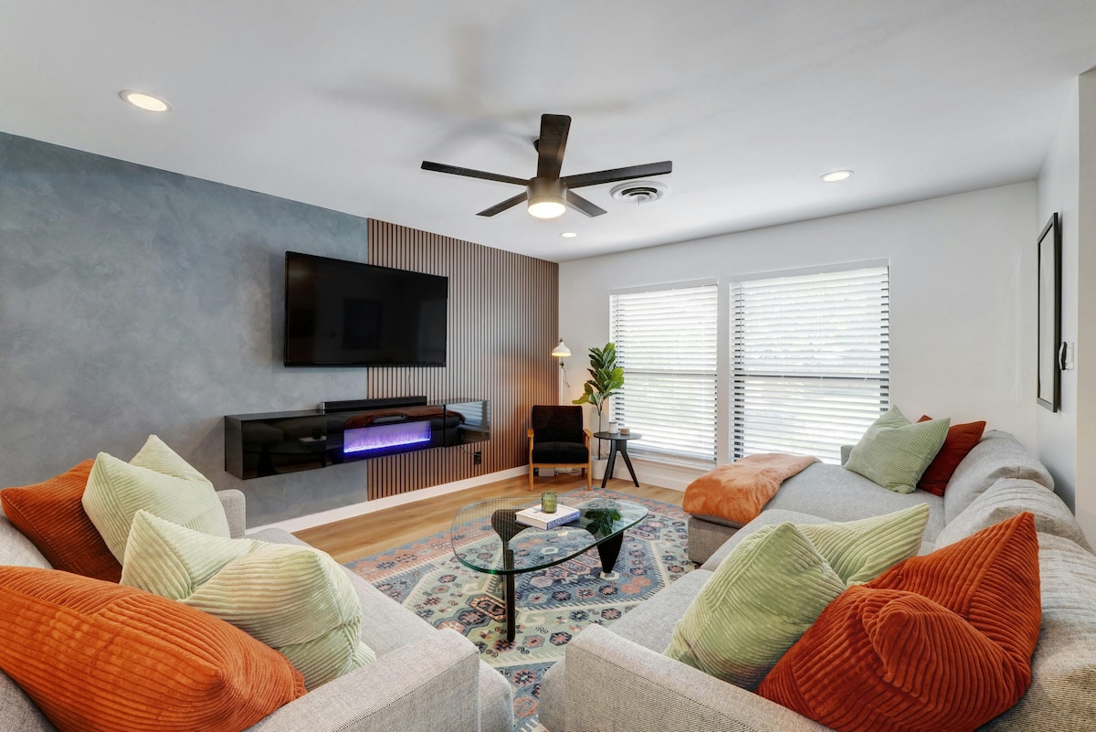 The living space features two comfortable sectional sofas adorned with vibrant throw pillows in shades of green and orange. A modern coffee table is centered on a large area rug, while a wall-mounted flat-screen TV and sleek entertainment unit add to the contemporary decor. Natural light filters through multiple windows.