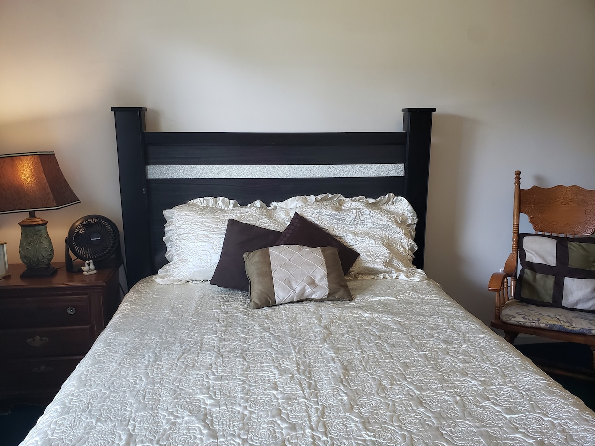 A bed with an intricate white bedspread is positioned against a plain wall. Decorative pillows in shades of brown and gold are arranged on the head of the bed. A bedside lamp and a wooden chair with a patchwork design are visible in the room.