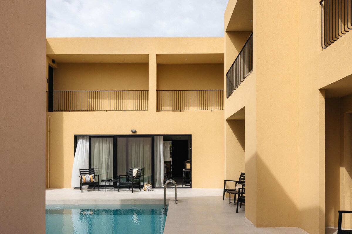 A tranquil pool area is framed by beige buildings with modern architecture. Comfortable outdoor seating is visible near the pool, offering a space for relaxation. Large glass doors reveal an inviting indoor area, seamlessly connecting the exterior and interior environments.