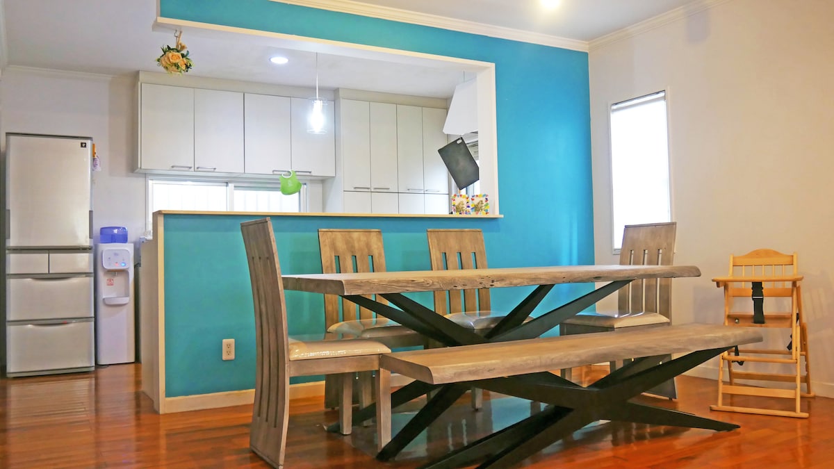 A dining area features a large wooden table with diagonal supports, surrounded by matching wooden chairs. A high chair is positioned at one end. The space is accentuated by a turquoise wall, and a kitchen area is visible in the background with modern cabinetry.