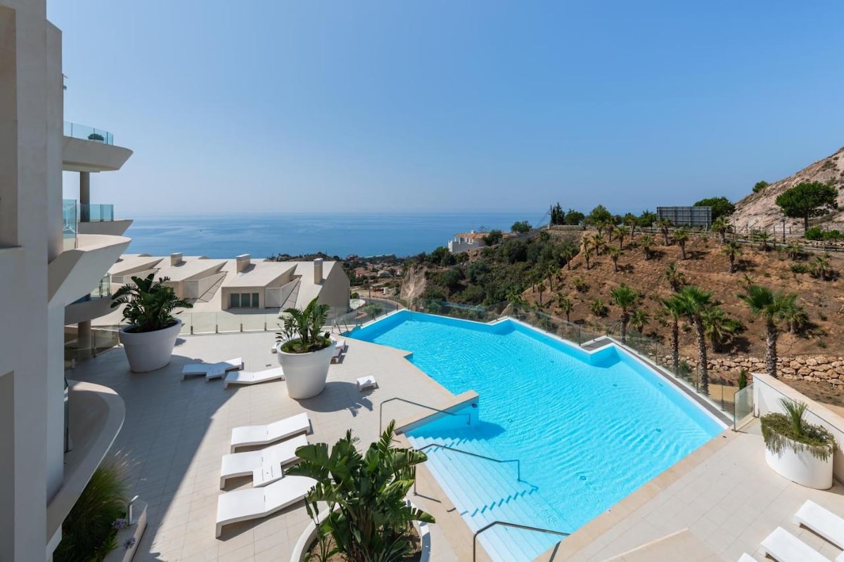 A large outdoor area features a tranquil swimming pool with steps leading into the water. Surrounding the pool, neutral-toned lounge chairs rest beside lush greenery and palm trees. The expansive view of the ocean and hills in the distance enhances the serene environment.