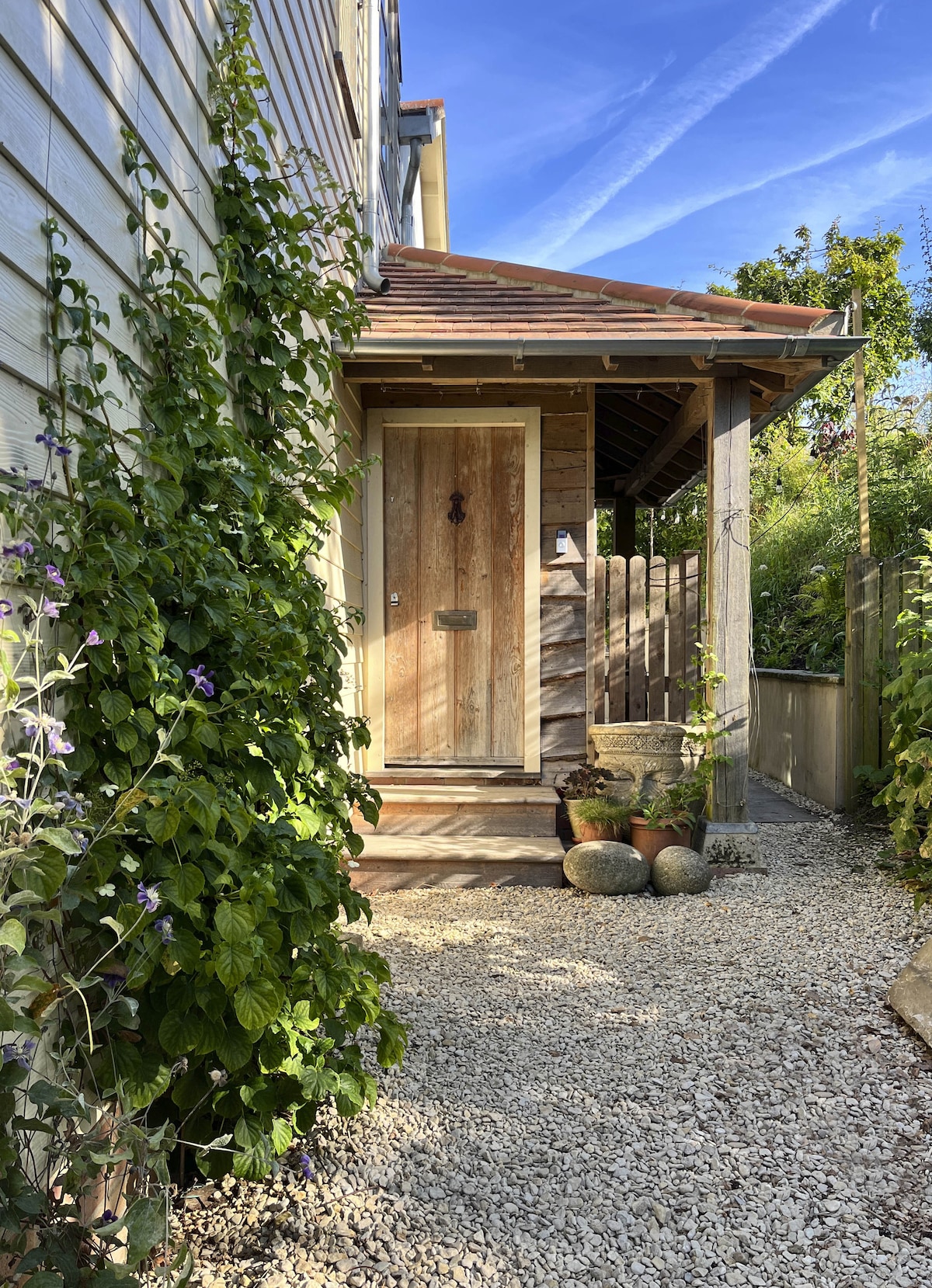 The entrance to the apartment is framed by lush greenery and flowered vines. A wooden door with a simple design is highlighted, complemented by a gravel pathway. Potted plants adorn the area, enhancing the welcoming ambiance of the entrance.