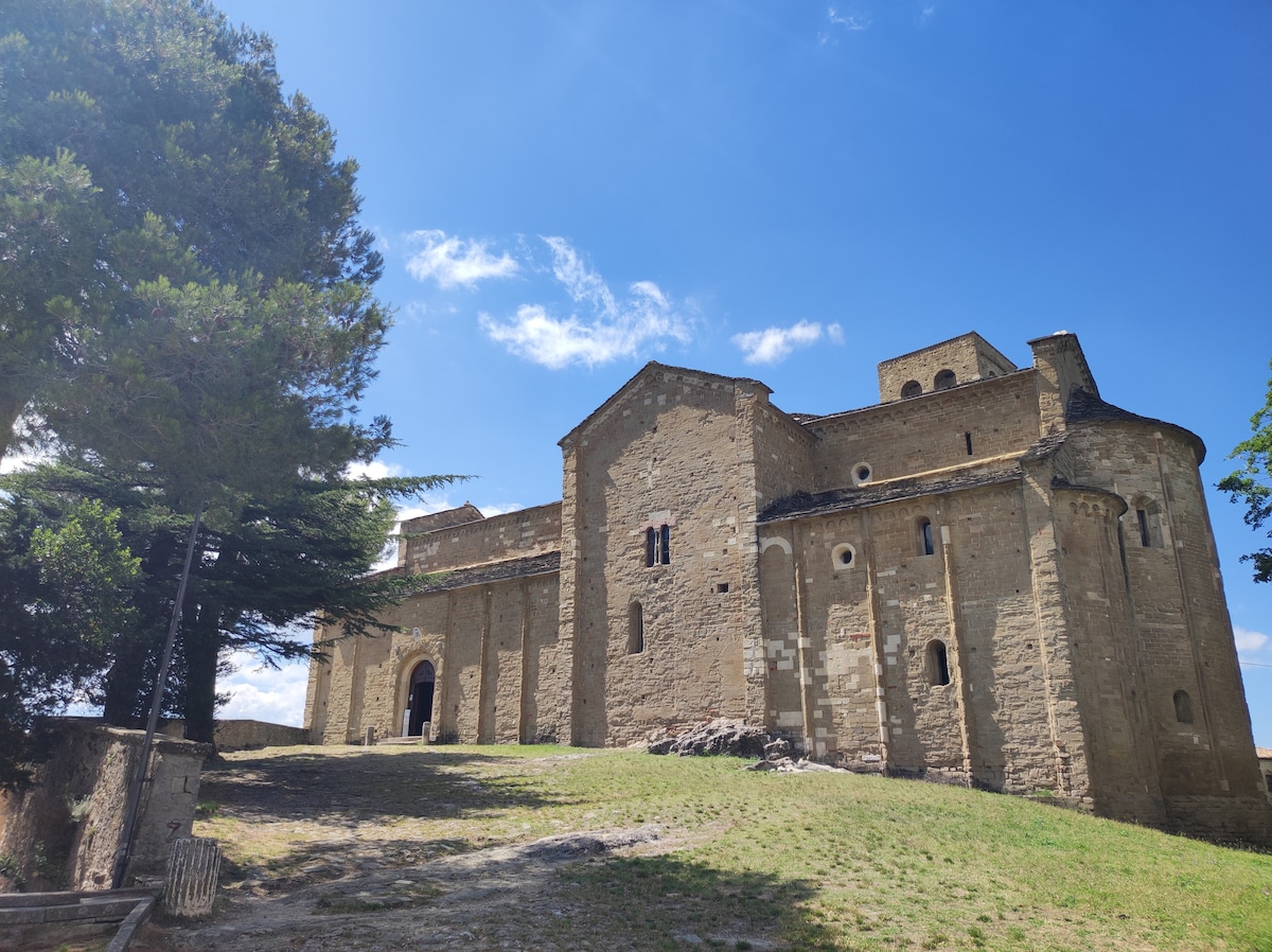An ancient stone building is depicted, showcasing a blend of architectural styles with a rugged exterior. The structure is surrounded by greenery and is set against a clear blue sky, providing a serene backdrop. The landscape descends gently, leading towards the entrance.