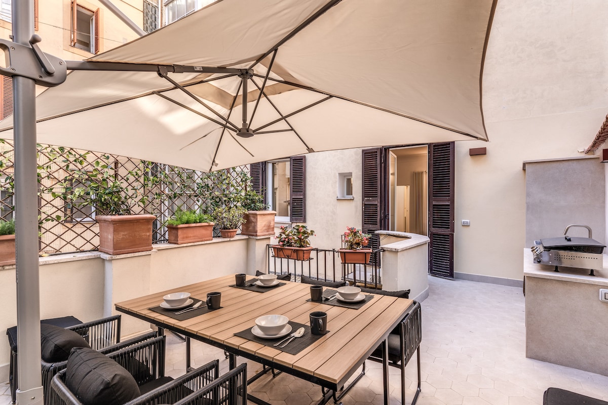 An outdoor terrace area features a large table with seating for dining. An umbrella provides shade, while potted plants add greenery. A barbecue grill is positioned nearby, and entry points to the apartment are visible in the background.