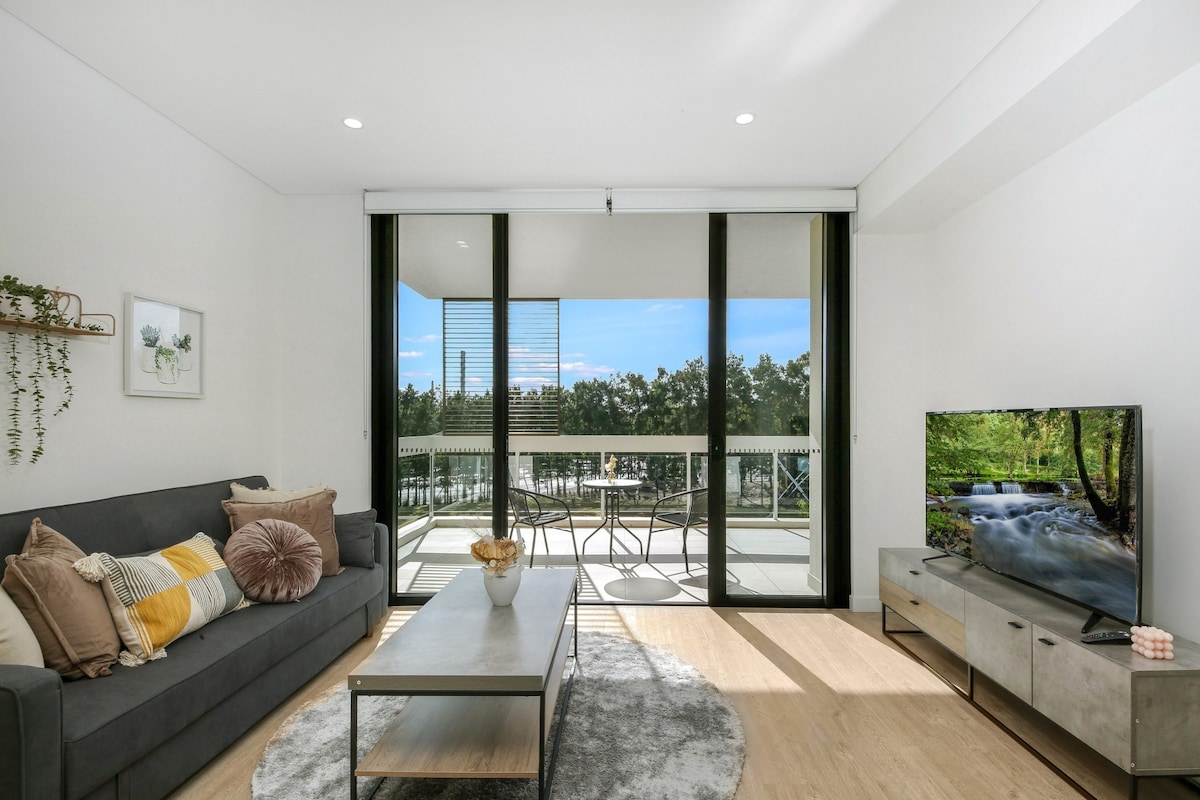 An open living area is showcased with a comfortable sofa adorned with decorative pillows. A coffee table sits in front, and large glass doors lead to a balcony overlooking greenery. A TV is positioned on a console against one wall, enhancing the modern design.