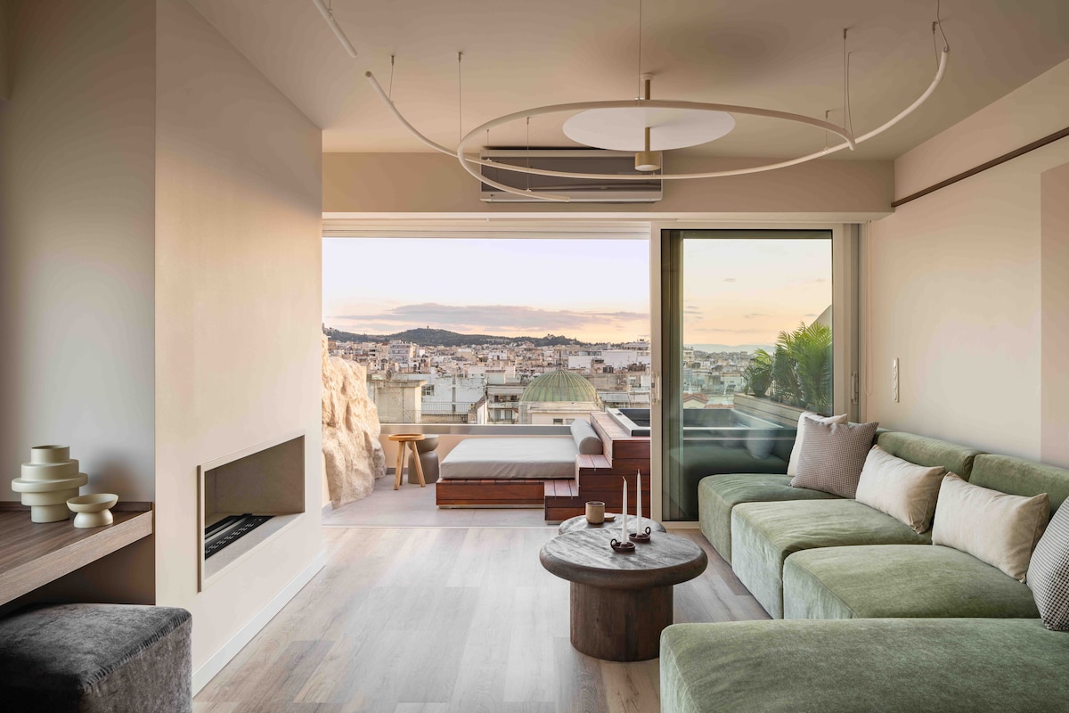 A modern living space features a plush green sofa and a circular coffee table. Large windows provide expansive views of the city and outdoor seating. Soft lighting is provided by a contemporary ceiling fixture, while a sleek fireplace adds an element of warmth.