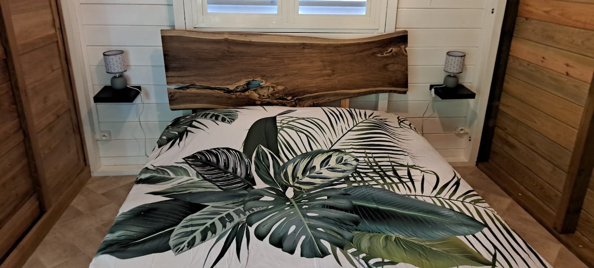 A spacious bed is adorned with a tropical-themed cover featuring lush green foliage. Unique wood paneling serves as a headboard, complemented by two bedside tables with lamps. Light is gently filtered through a window, enhancing the natural ambiance of the room.