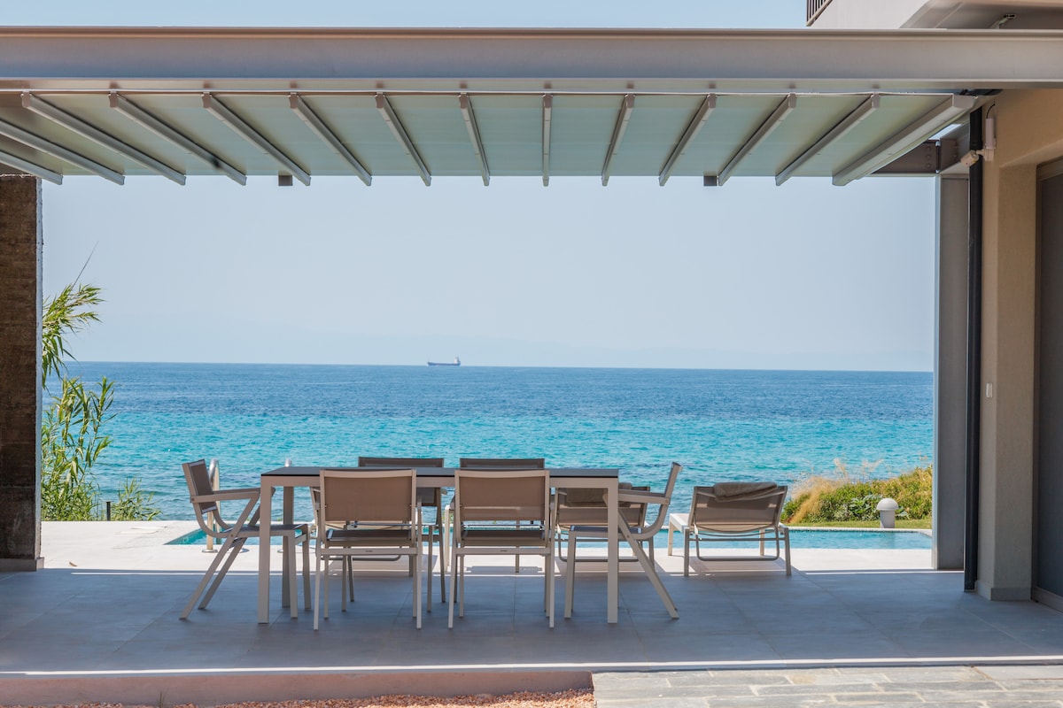 An outdoor dining area is furnished with a large table and multiple chairs, set beneath an awning. The view includes a calm sea with a distant ship and a pool in the background, framed by green landscaping.