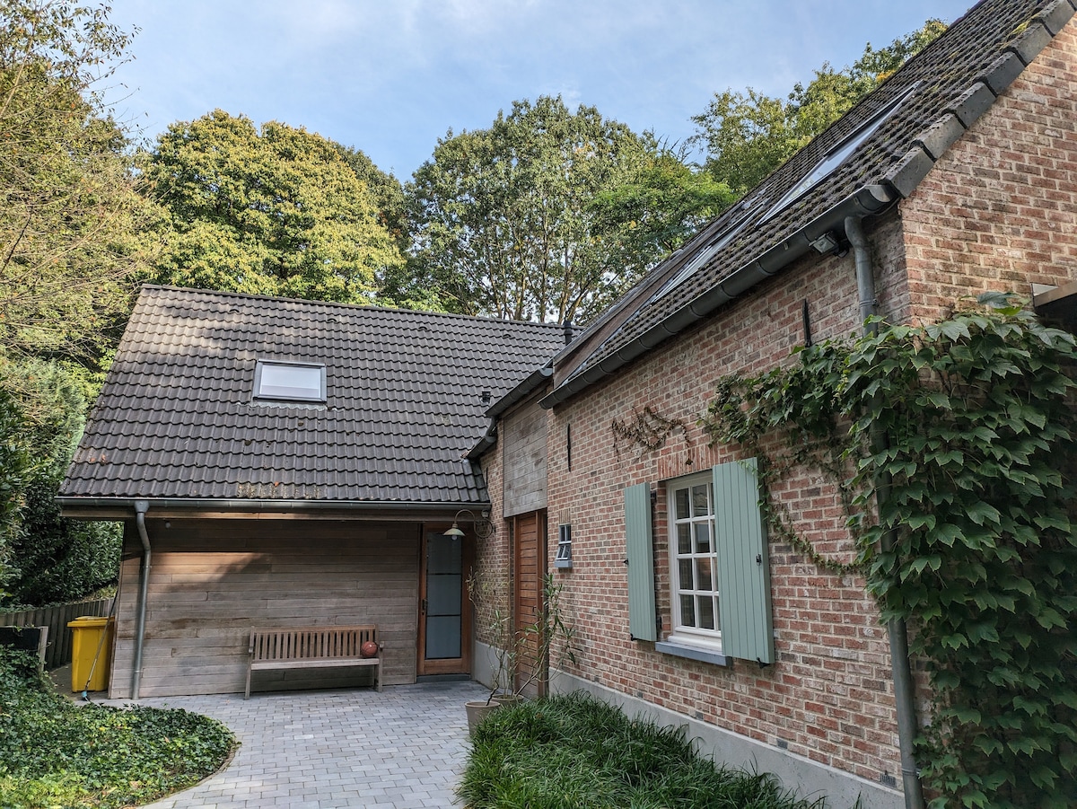 The exterior of a charming home features a mix of brick and wooden elements, surrounded by greenery. A welcoming entrance is highlighted by a bench, while a sloped roof and shutters add character to the facade. The paved pathway leads to the inviting front door.