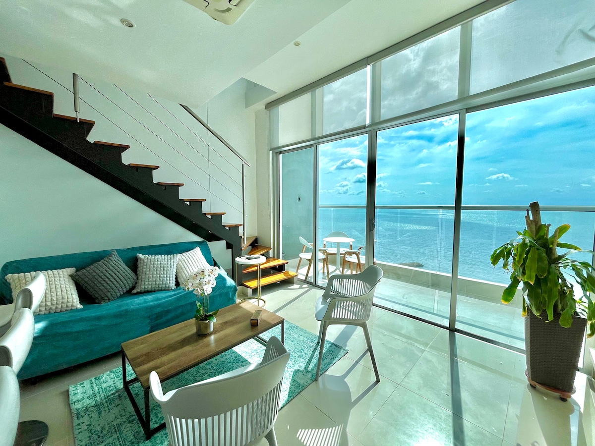 A spacious living area features large glass doors that provide an unobstructed view of the sea. A comfortable turquoise sofa is accompanied by white chairs surrounding a wooden table. Natural light fills the room, highlighting the elegantly placed indoor plants.