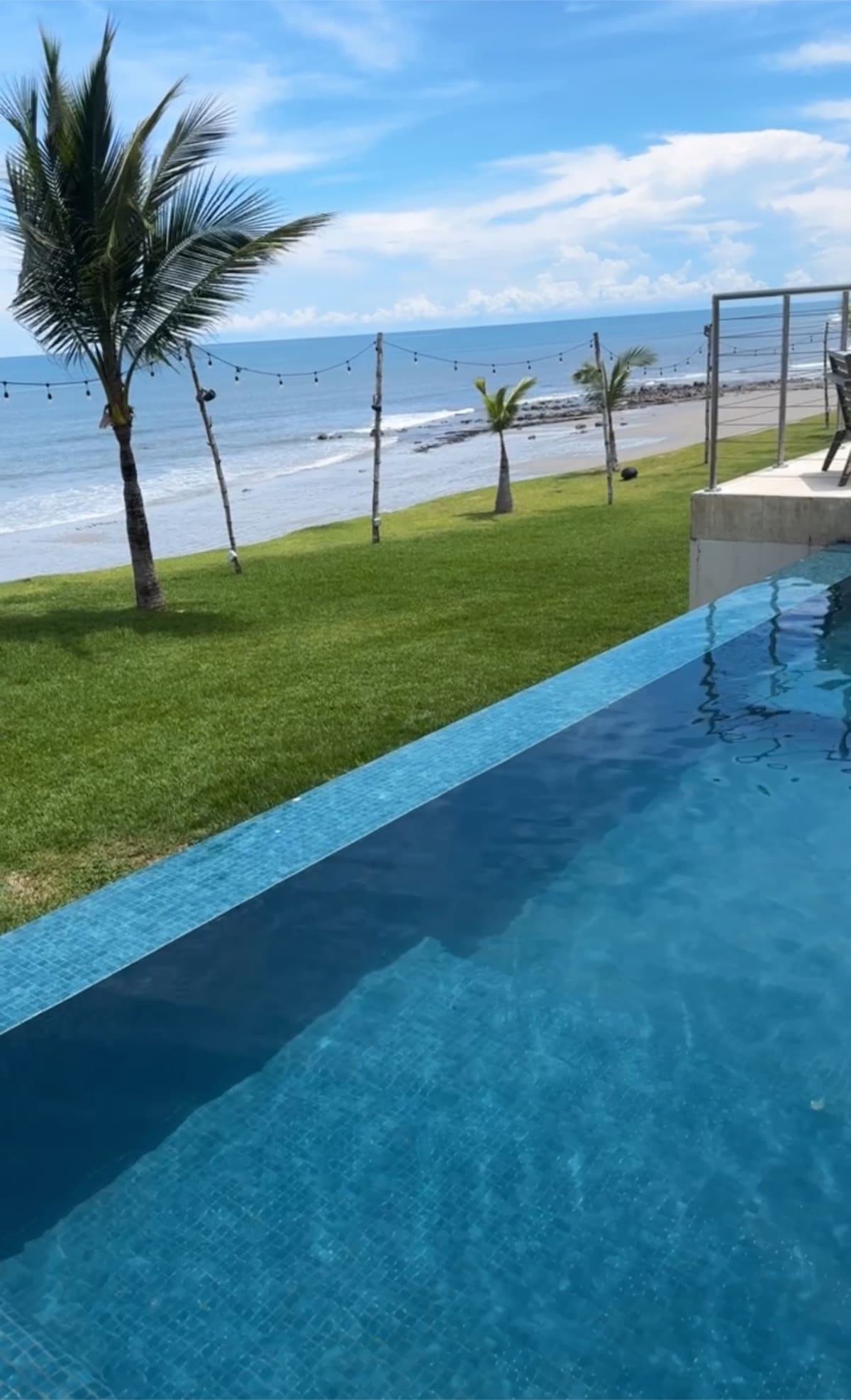 A tranquil oceanfront scene features a shimmering pool reflecting sunlight, with green grass and palm trees framing the view. The beach is visible beyond the water, along with a clear blue sky adorned with scattered clouds.