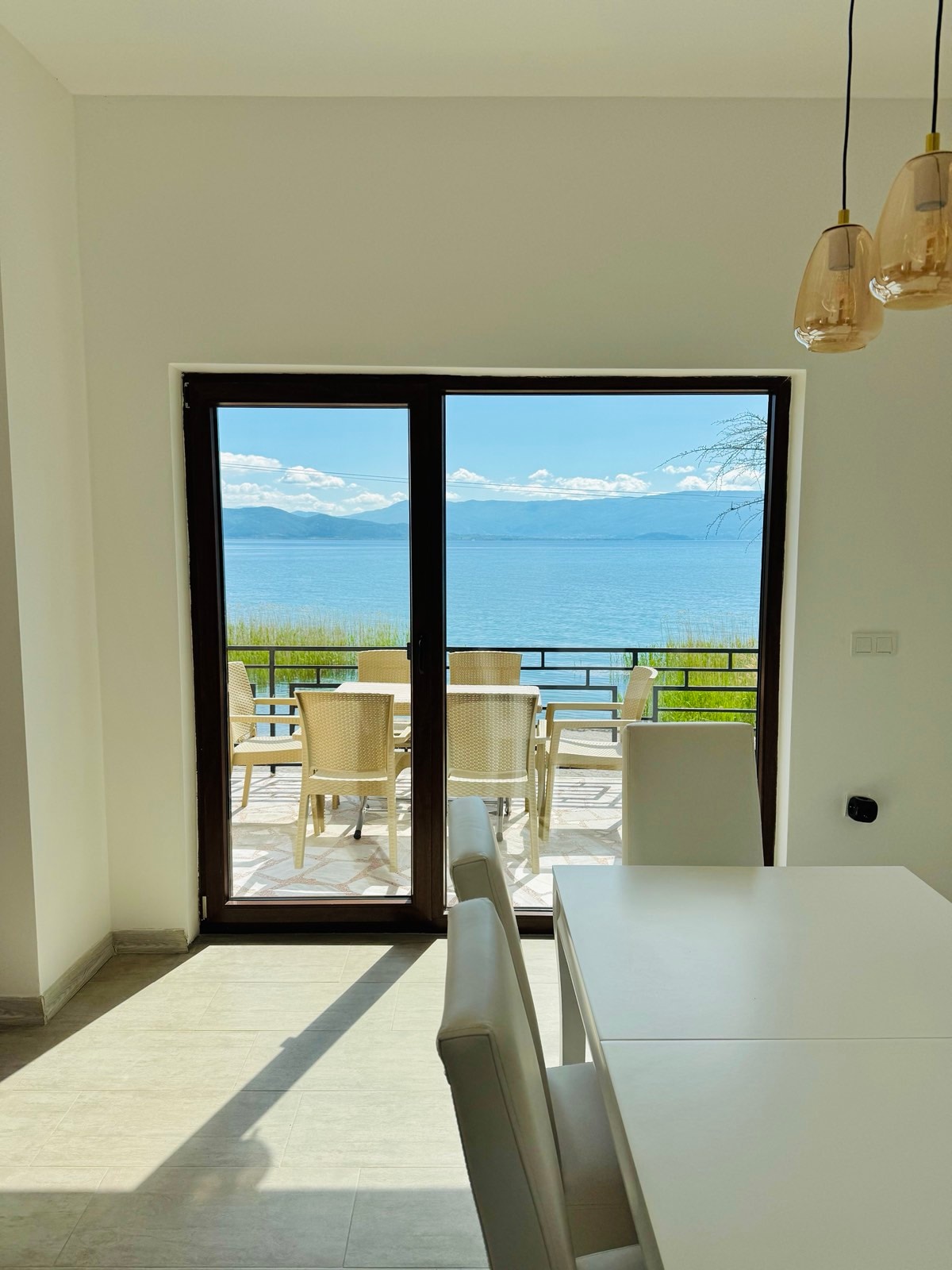A view from inside the villa reveals a glass door leading to a spacious terrace with outdoor seating. Natural light fills the room, highlighting a dining table. The serene lake and distant mountains are visible through the door, creating a calming backdrop.