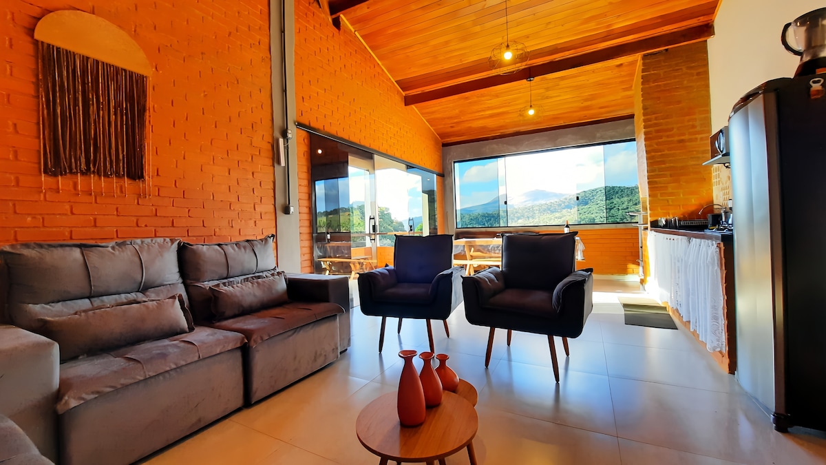A open living area is highlighted by large glass windows that offer views of the surrounding mountains. Comfortable seating includes a gray sofa and two dark chairs arranged around a small wooden table, which is complemented by terracotta vases.