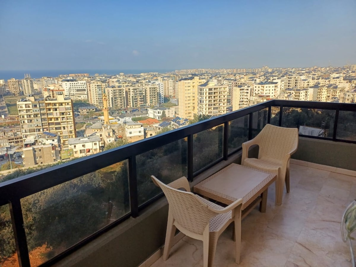A balcony offers outdoor seating with two light-colored chairs and a small table, overlooking a panoramic city view. The horizon features a blend of urban architecture and distant coastal scenery under a clear blue sky.