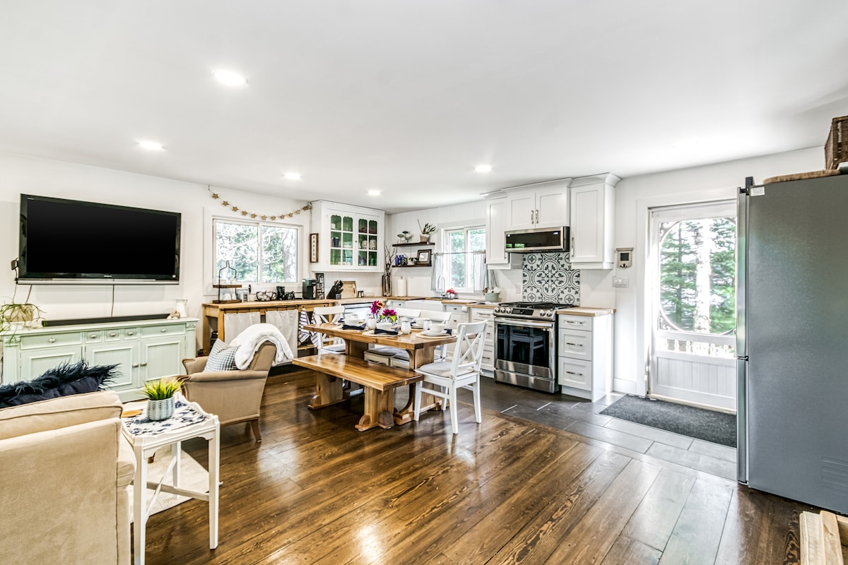The open-plan living space features a large dining table surrounded by comfortable seating. A cozy sitting area includes a television mounted on the wall. Natural light fills the room from multiple windows, highlighting kitchen appliances and cabinetry.