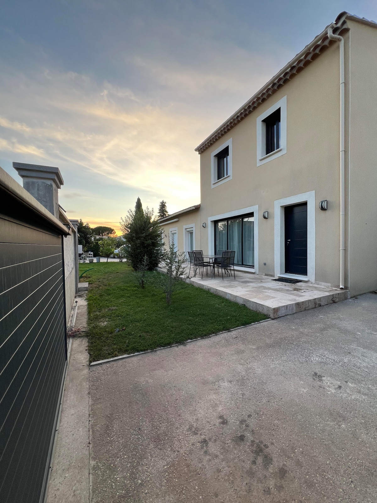 The exterior of the villa showcases a modern design with large windows and a terrace furnished with dining chairs. A small green lawn complements the exterior, and the setting sun casts a warm light over the scene, enhancing the peaceful atmosphere.