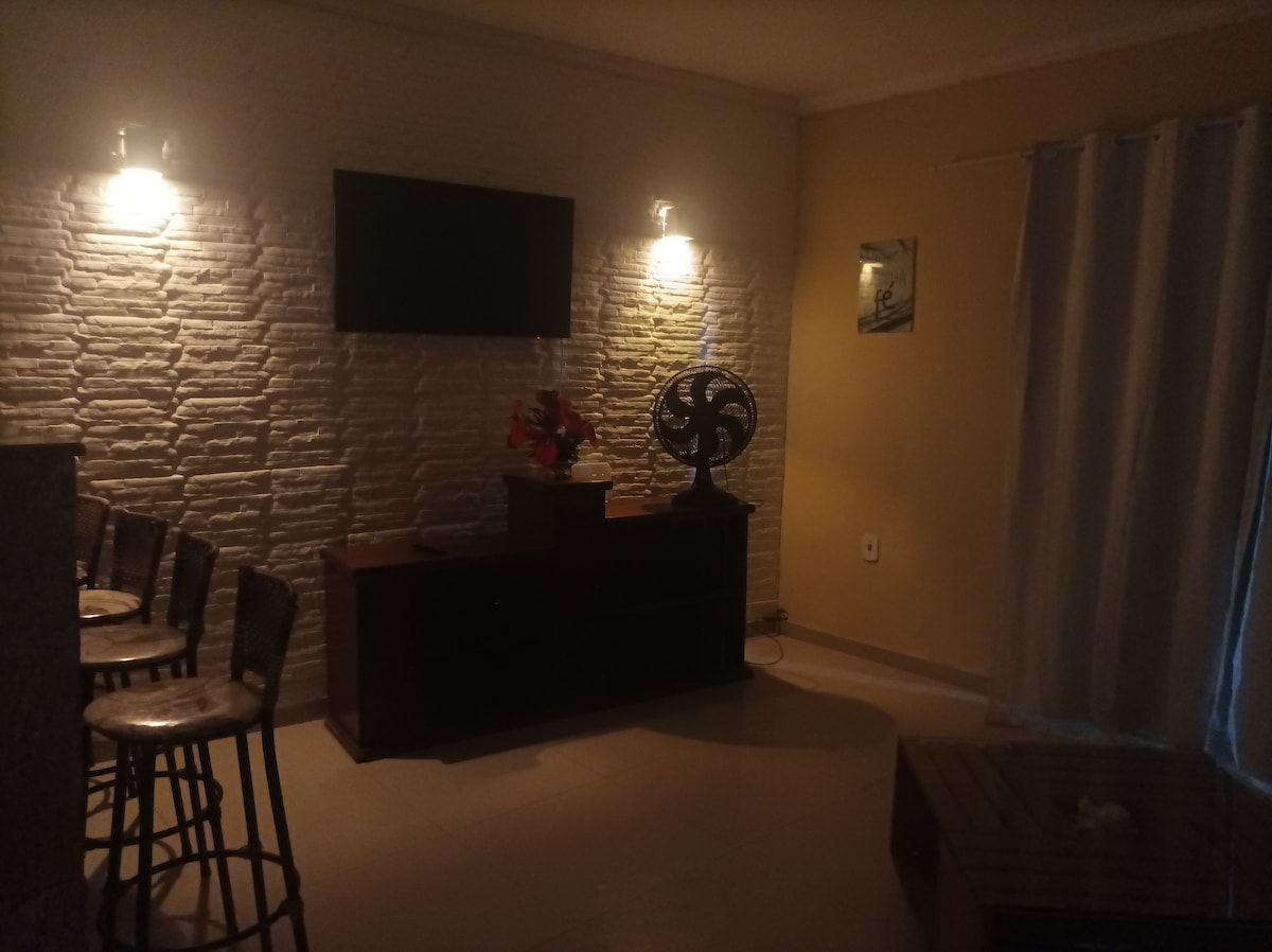 A living area is featured with a flat-screen television mounted on a textured wall. Two wall sconces provide soft lighting. A wooden console table sits beneath the TV, accompanied by a decorative plant. A fan and a small coffee table enhance the space's functionality.