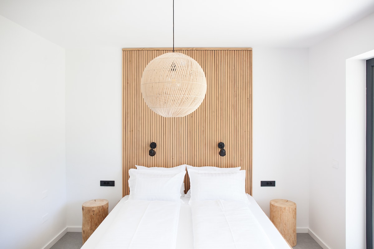 A modern bedroom features a light wood accent wall behind a spacious bed with two separate mattresses. Natural light fills the room through a large window, and two minimalist bedside tables are placed on either side. Stylish pendant lighting adds an elegant touch.