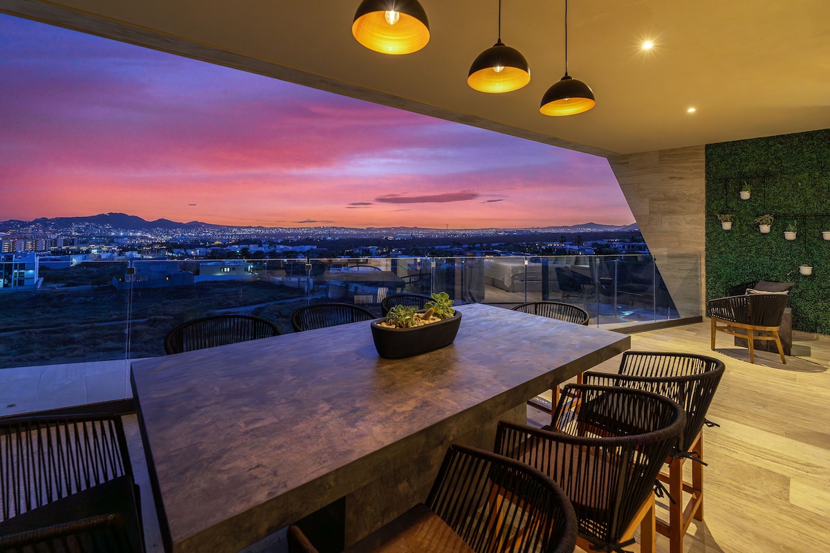 An outdoor dining area is featured on a spacious terrace, showcasing a large table surrounded by elegant chairs. Soft lighting from pendant fixtures illuminates the space, which overlooks a vibrant sunset sky. A lush green wall adds a natural touch to the elegant setting.