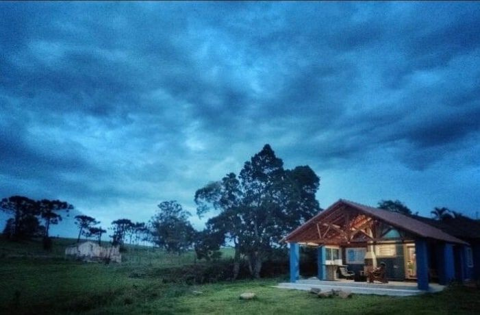 A mountain house is positioned amidst a lush green landscape, under a cloudy blue sky. Its exterior features a rustic charm, with a spacious porch visible. Large windows allow light to enter, suggesting a cozy atmosphere. Surrounding trees add to the natural setting.