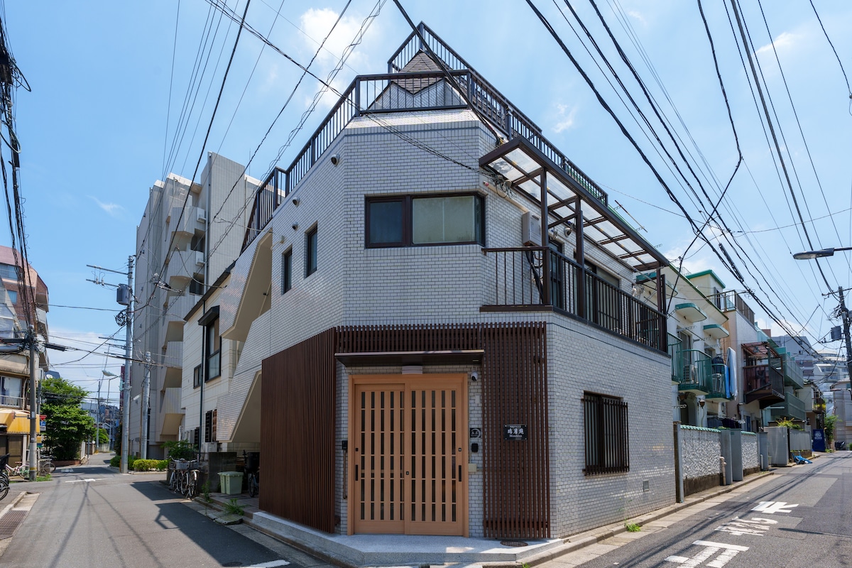 The exterior of a multi-story building is presented, featuring a mix of white brick and wooden paneling. Large windows allow for natural light, while a rooftop terrace is visible. The street is lined with power lines and nearby structures, conveying an urban environment.