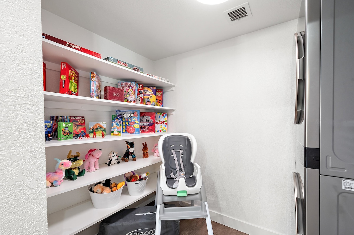 A well-organized storage area features shelves filled with colorful toys and games, suitable for children. A high chair is positioned nearby, and a cozy assortment of stuffed animals complements the playful atmosphere. The space maintains a clean and inviting appearance.