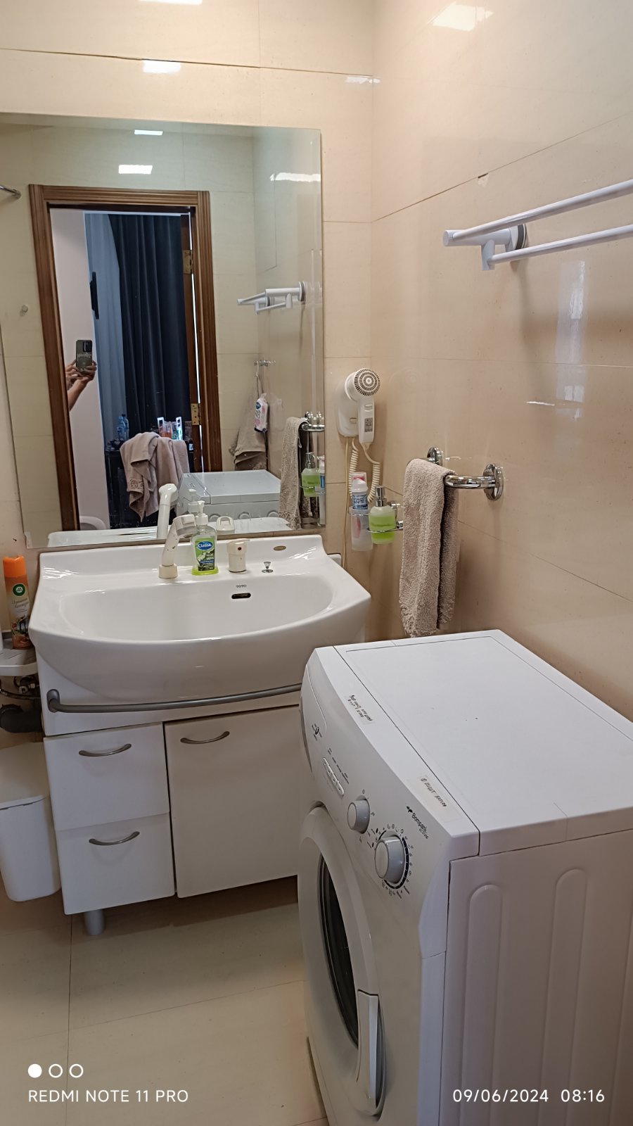 The bathroom features a modern sink with an adjacent mirror and several toiletries neatly arranged. A washing machine is positioned beside the sink, and a towel hangs on the chrome towel bar. Soft lighting enhances the clean, minimalistic design.