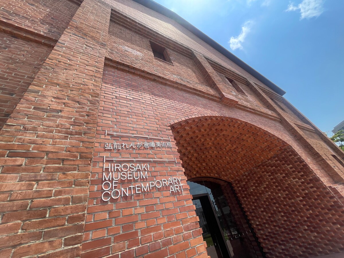 The entrance of the Hirosaki Museum of Contemporary Art is showcased, featuring distinctive red brick architecture. The archway is prominent, with the museum's name displayed in both English and Japanese. Surrounding greenery enhances the inviting appearance of the building.