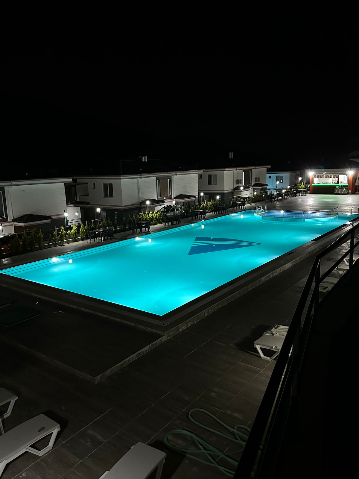 A well-lit outdoor pool area is featured, displaying turquoise water illuminated by soft, ambient lighting. Surrounding the pool are lounge chairs arranged neatly, creating a relaxing environment for guests. The scene is set at night, enhancing the tranquil atmosphere of the space.