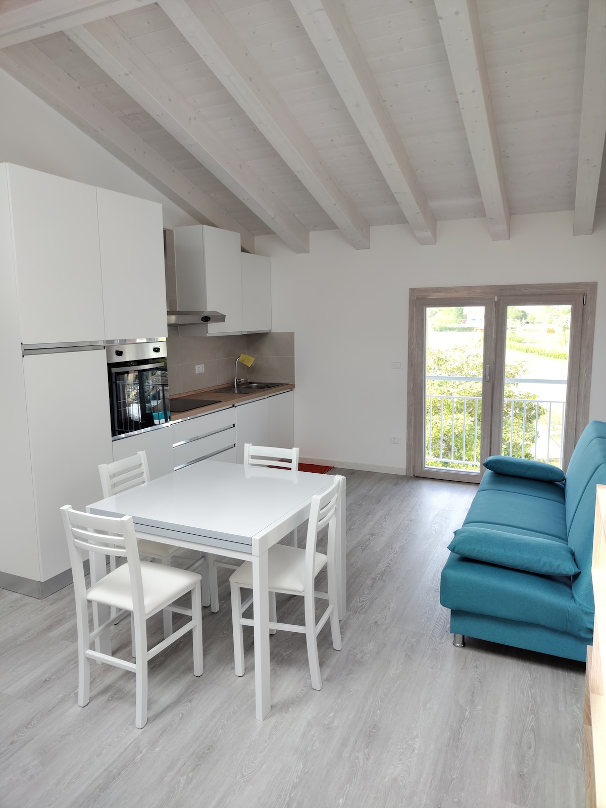 The bright kitchen area features modern cabinetry and appliances, including an oven and refrigerator. A dining table with four white chairs is positioned in the center. A turquoise sofa complements the space, with a view offered through glass doors leading to a small balcony.