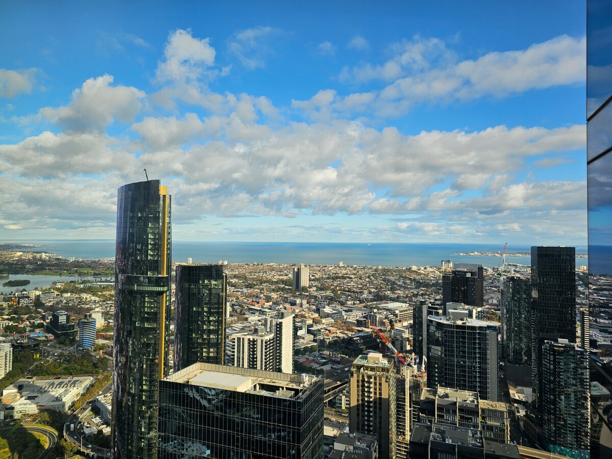 A panoramic view captures Melbourne’s skyline from the 65th floor, featuring modern buildings and the distant coastline. The expansive sky showcases varied cloud formations above the urban landscape, with the shimmering waters of the bay visible on the horizon.