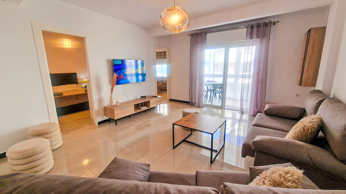 A bright living room features comfortable seating arranged around a low coffee table. A wall-mounted television is visible alongside large glass doors that lead to a balcony. Light-colored walls and smooth flooring enhance the spacious feel, while sheer curtains allow for natural light.