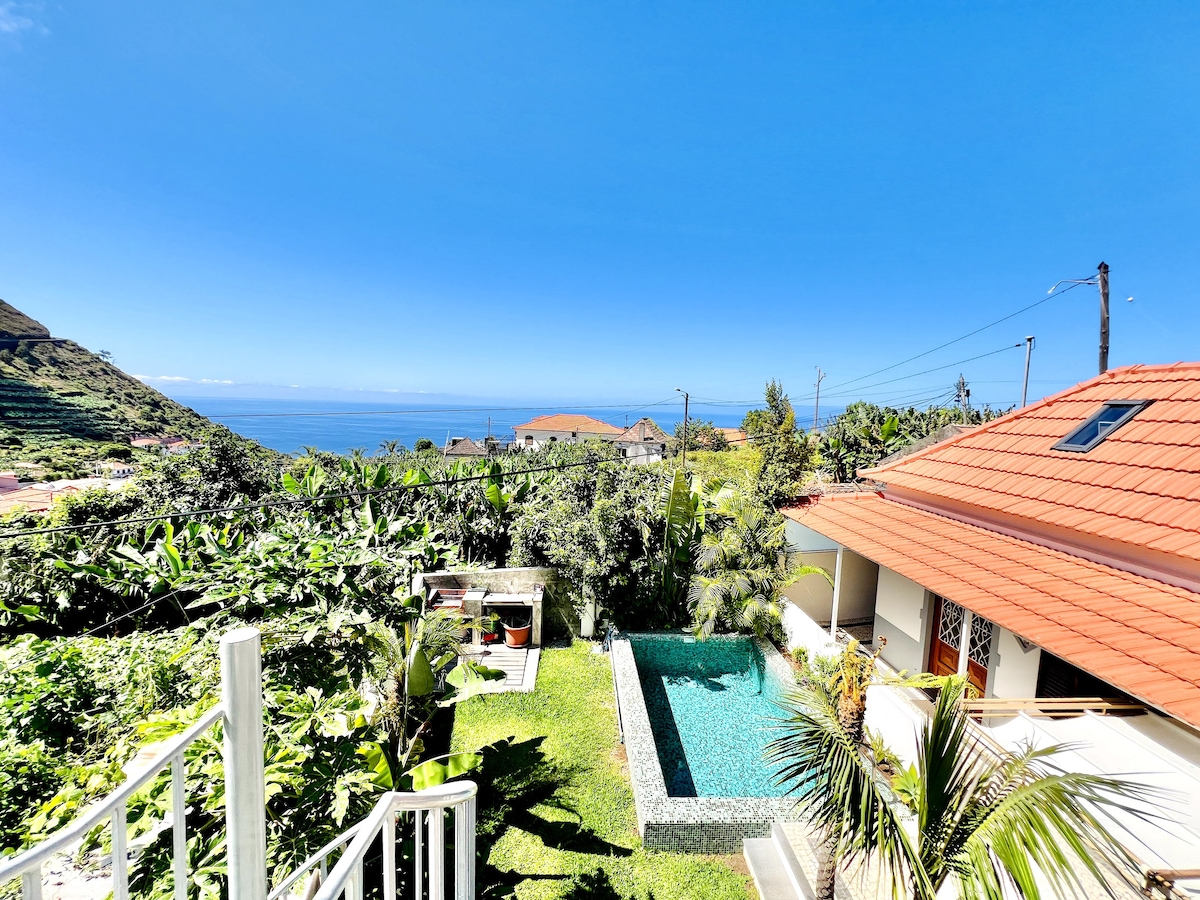An expansive view captures a tranquil pool set within a landscaped garden, flanked by lush banana plantations. The sea extends towards the horizon, while a section of the house, featuring a red-tiled roof, can be seen in the foreground.
