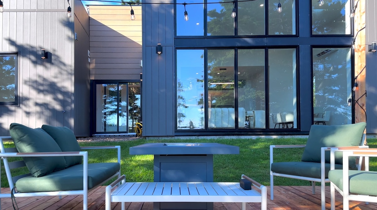 An outdoor seating area features modern furniture positioned on a wooden deck. Two armchairs with cushions are complemented by a low coffee table. Large windows of the house reflect natural light and provide a view of the green lawn and trees surrounding the property.