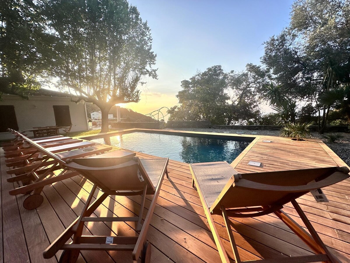 A wooden deck surrounds a heated swimming pool, with multiple loungers positioned for relaxation. The serene landscape features lush trees, and the soft glow of sunset illuminates the scene, creating a tranquil atmosphere ideal for unwinding.