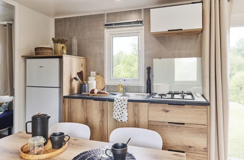 A modern kitchen features wood cabinetry and a compact dining area. Stainless steel appliances, including a refrigerator and a stovetop, are visible. Natural light enters through a nearby window, highlighting a table set with two mugs and a jar, creating a functional cooking and dining space.