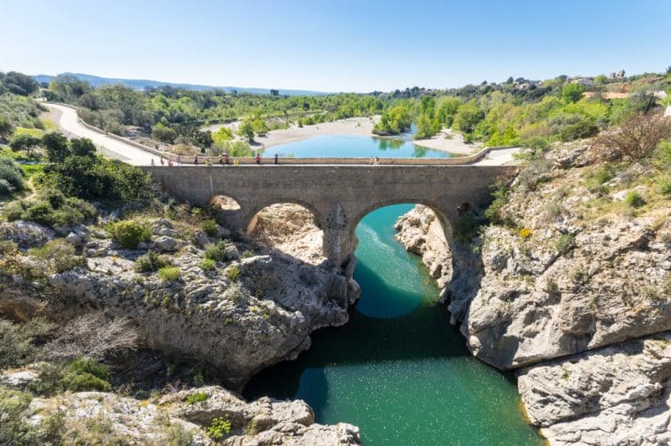 An arched stone bridge spans a serene body of water, surrounded by lush greenery and rocky terrain. Clear blue water reflects the landscape, while people can be seen walking along the pathway above the bridge, emphasizing the tranquil setting.