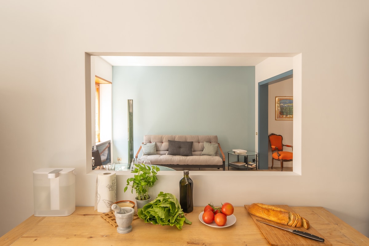 A view from the kitchen features fresh ingredients like lettuce, tomatoes, and a loaf of bread on a wooden table. Through a pass-through opening, a cozy living area is seen, showcasing a light-colored sofa against a soft blue wall.