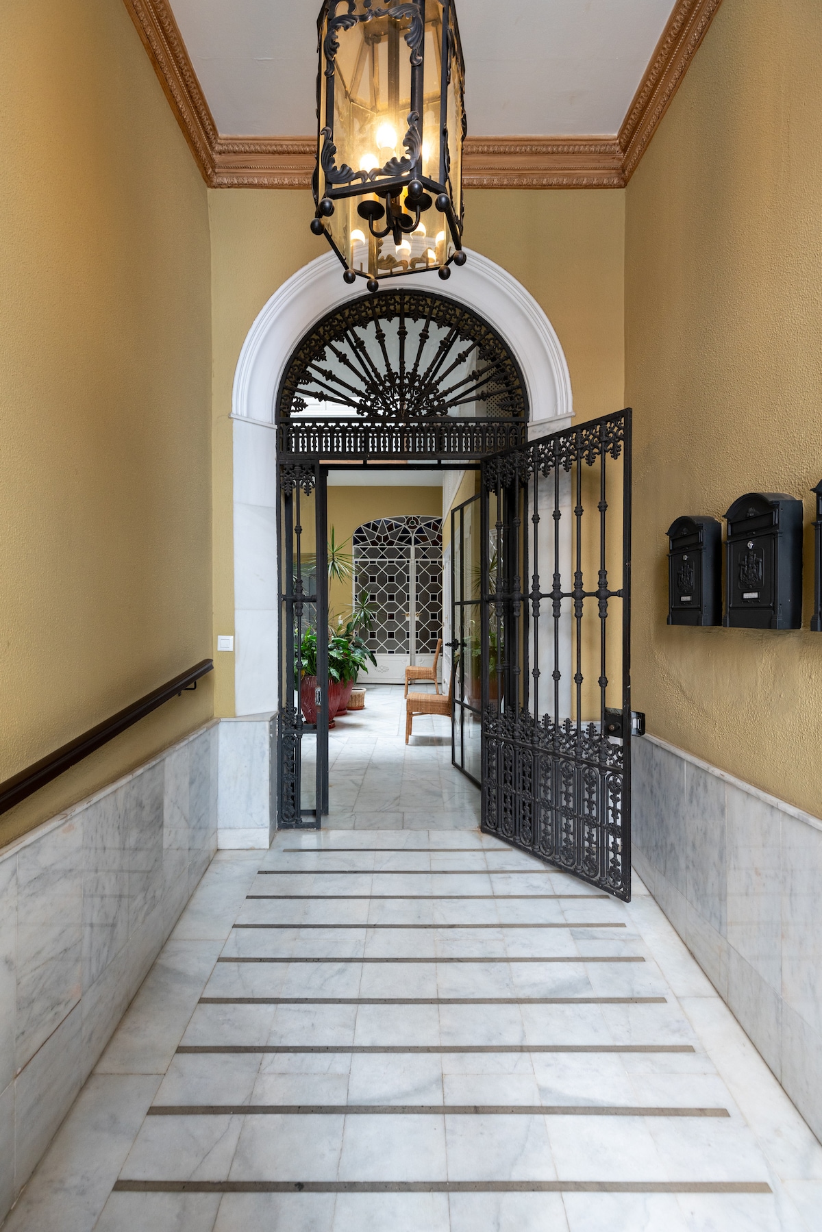 A grand entrance is depicted with a wrought-iron gate opening to a tiled hallway. Marble flooring is accentuated by subtle lines, while potted plants and bright natural light create an inviting ambiance. Architectural details include an arched doorway and an ornate ceiling fixture.