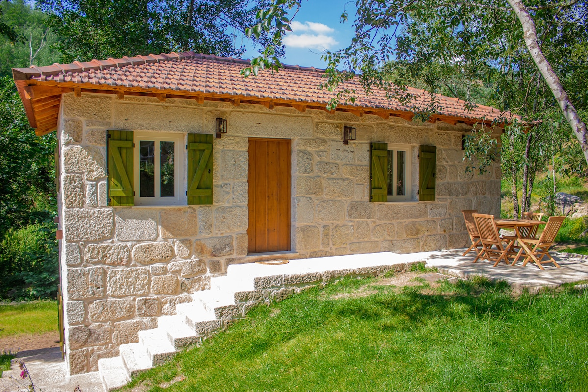 A charming stone cottage is framed by a lush green landscape. The structure features wooden shutters in a vibrant green, a warm wooden door, and a front porch. A small outdoor seating area with two chairs and a table is positioned on the grassy lawn.