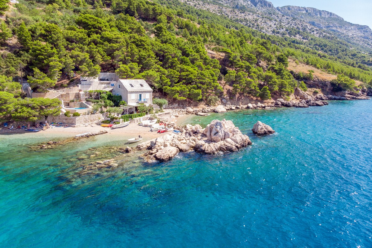 The image captures a serene coastal scene featuring a traditional structure nestled among lush greenery. Crystal-clear waters gently lap against rocky outcrops, with a sandy beach visible below. The surrounding landscape displays dense pine trees and rugged hills, enhancing the natural beauty of the location.