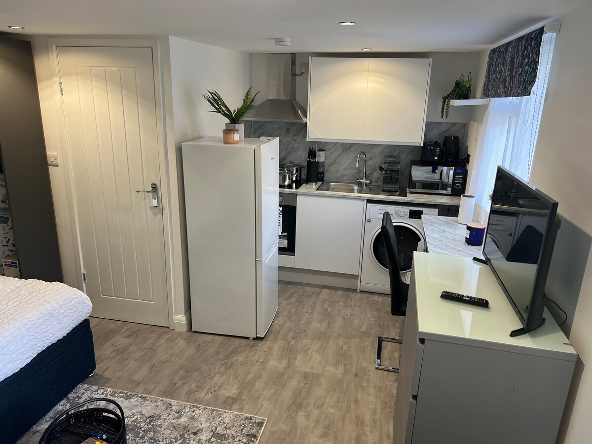 A well-equipped studio kitchen features modern appliances, including a fridge, microwave, and washing machine, with ample countertop space. A dining area is present, complemented by a TV mounted on a light-colored cabinet. Soft lighting enhances the functional layout of the space.