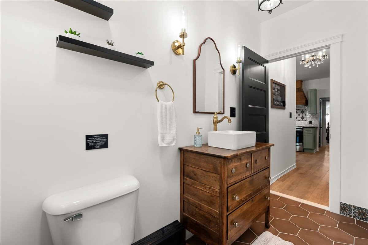 A modern bathroom is presented with a wall-mounted shelf displaying small plants. A stylish wooden dresser holds a basin sink with a gold faucet. A large mirror reflects simple decor, while a white towel hangs nearby. Elegant light fixtures enhance the room's ambiance.