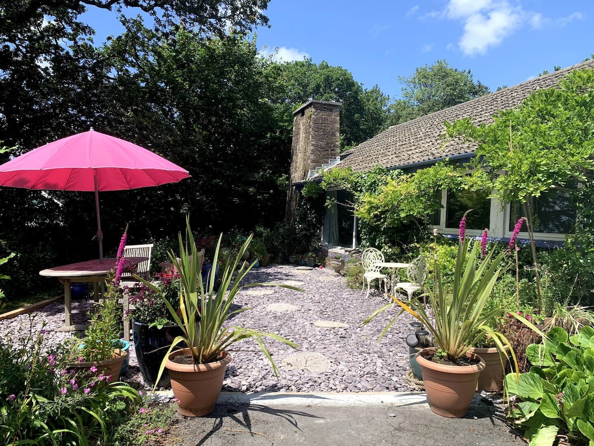 The exterior garden area features lush greenery and colorful flowers surrounding a gravel path. A pink umbrella provides shade over a round table with chairs. Potted plants add vibrancy, while a stone chimney is visible on the building, enhancing the natural setting.