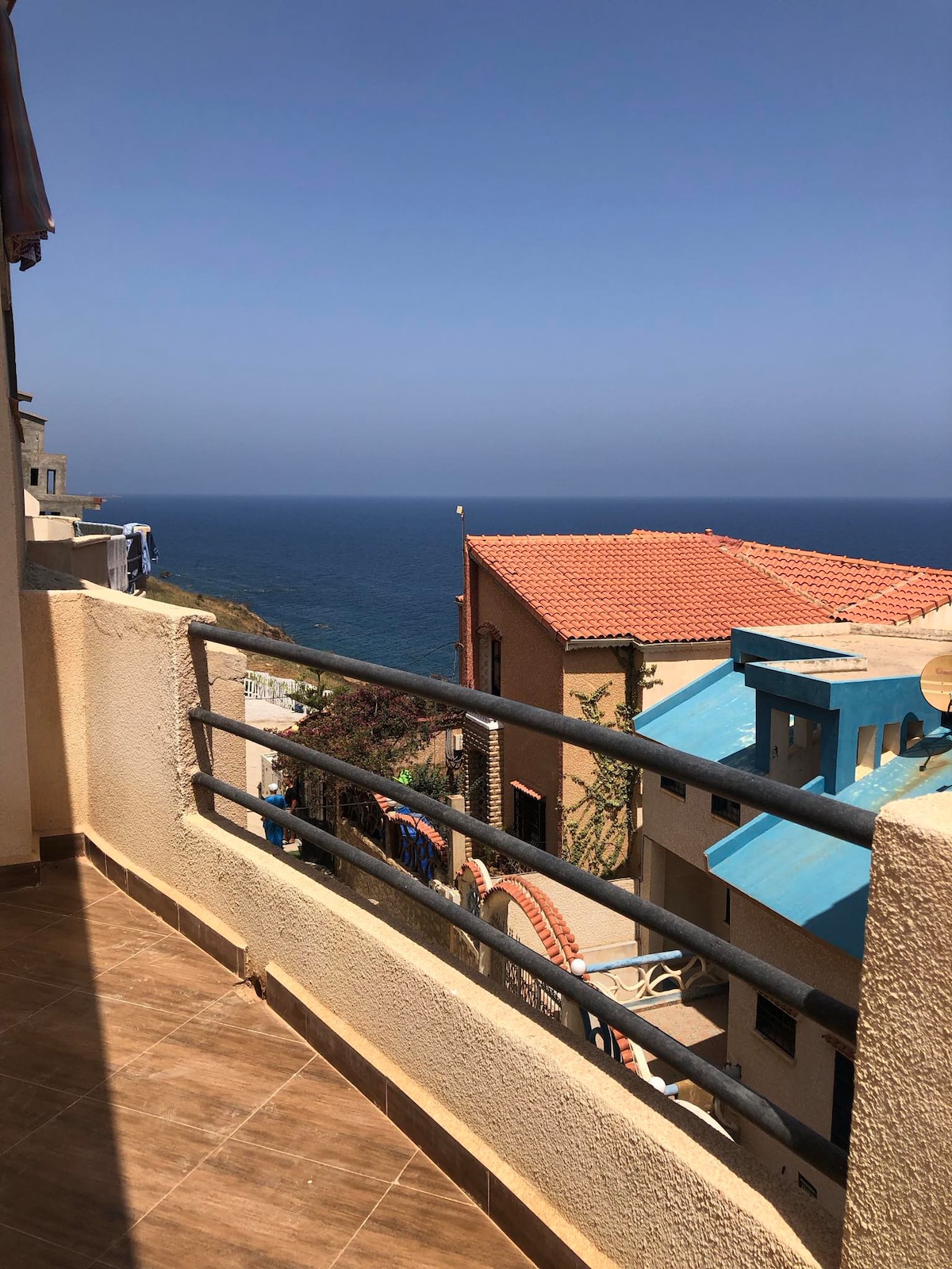 A balcony space is visible, featuring a railing with a view of a tranquil blue sea extending toward the horizon. Nearby buildings display tiled roofs and colorful accents, while the sunny sky contributes to a bright atmosphere.