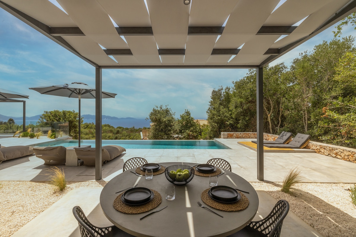An outdoor dining area features a round table set with dark plates and transparent glasses, surrounded by elegant black chairs. Sun loungers rest beside a pool, with a backdrop of lush greenery and distant mountains, while large umbrellas provide shade.