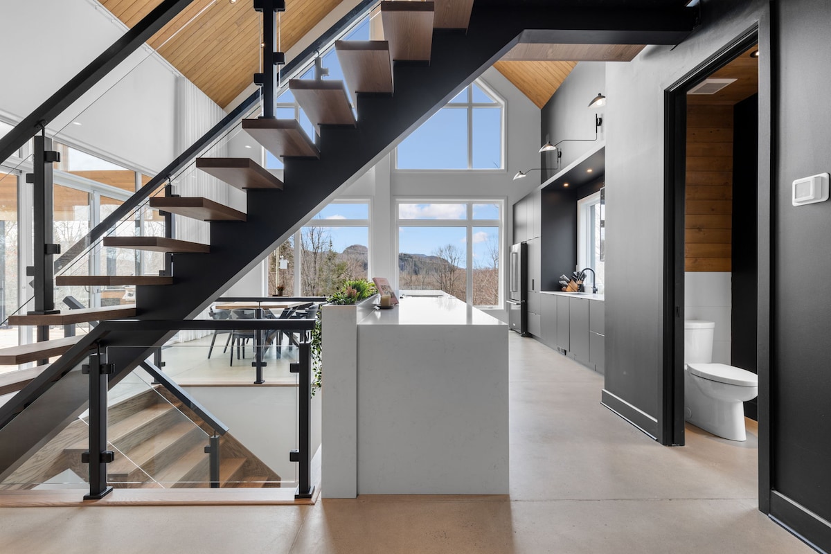 A modern open-concept space features a staircase with a sleek metal railing, leading to upper levels. Large windows allow natural light to fill the area, while views of the surrounding landscape are visible. The kitchen and dining space are organized and stylishly designed.